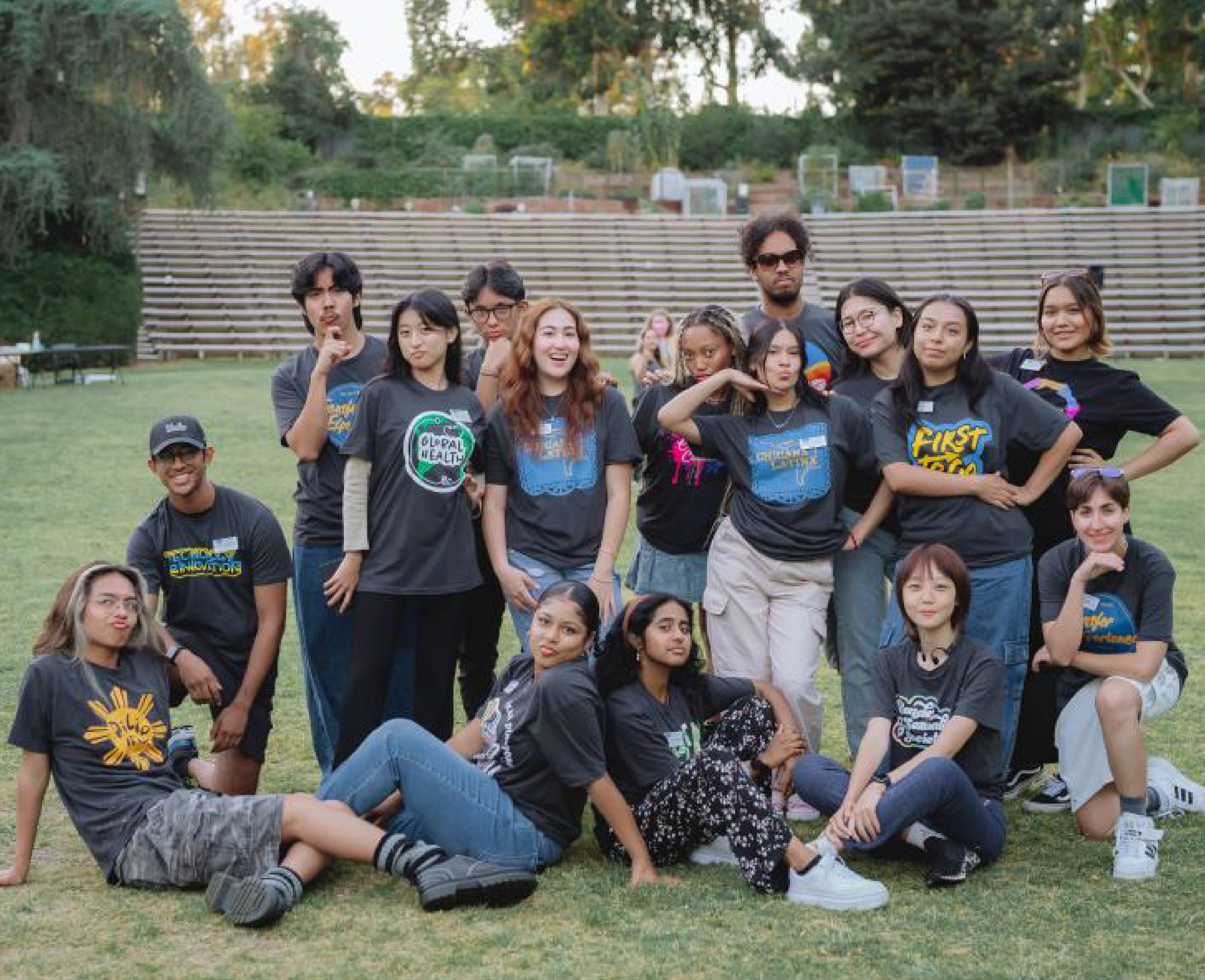 Una foto de grupo tomada al aire libre de diversos estudiantes, todos con varias camisetas gráficas, de pie y sentados juntos en un campo de césped. Los estudiantes sonríen y posan alegremente, creando una sensación de camaradería.