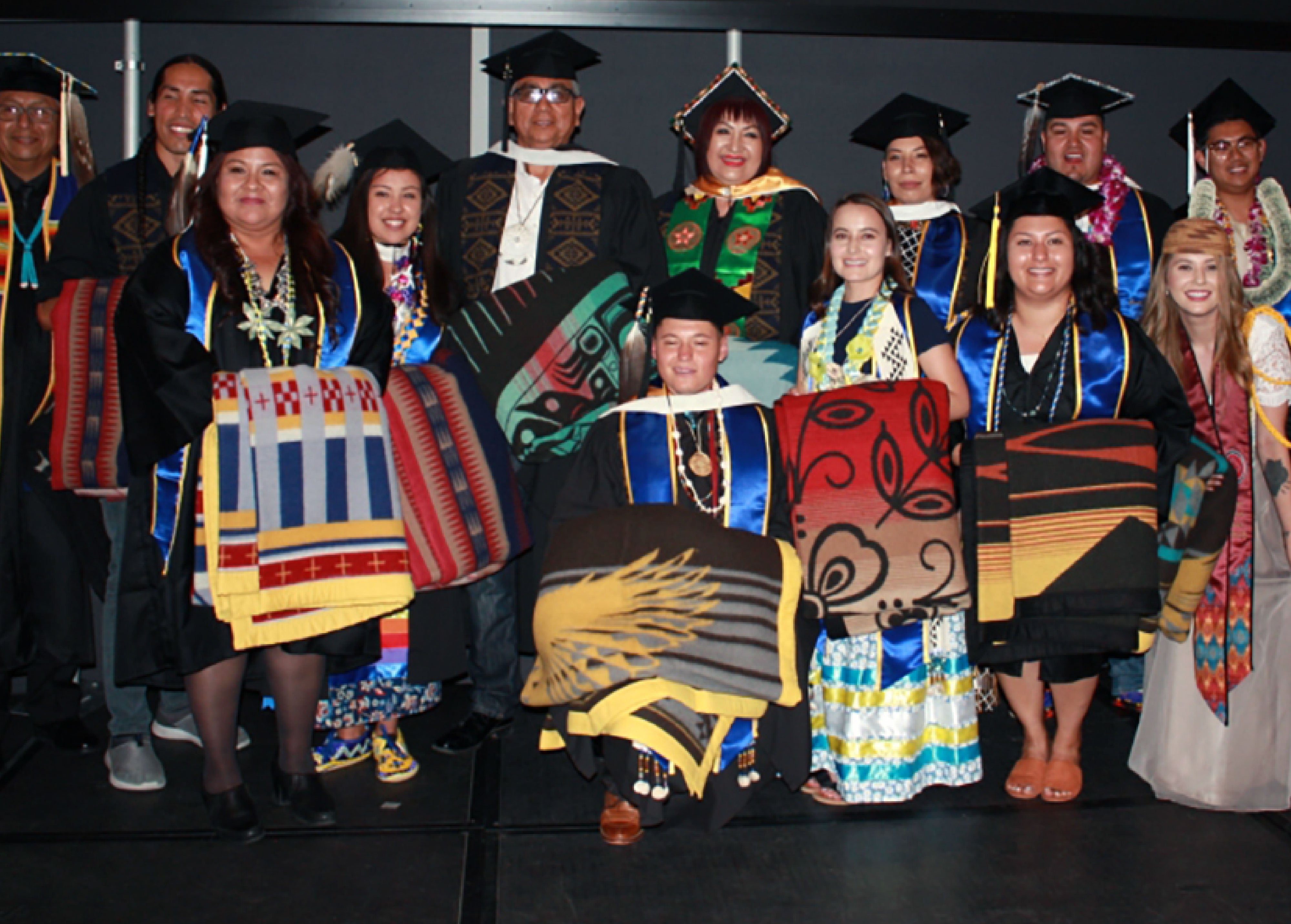 Group of UCLA graduates from the American Indian Studies program proudly posing in their caps and gowns. They are wrapped in vibrant, traditional Native American blankets, symbolizing cultural heritage and academic achievement. Each person displays a unique blanket design, adding to the richness of the celebration, while they stand together as a diverse representation of Native American cultures and traditions.