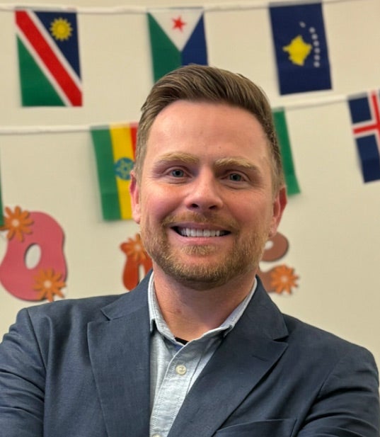 Andrew McClure smiles while wearing a gray blazer and light blue shirt. Colorful flags and decorations are displayed in the background.