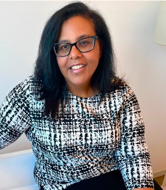 Anya Booker smiles while wearing a black-and-white patterned top. She is seated against a neutral background.