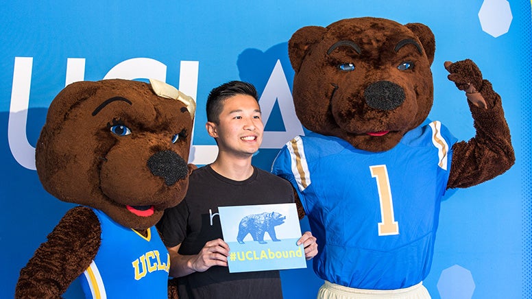 A student holding a #UCLAbound sign smiles as he poses with UCLA mascots Joe and Josie Bruin.
