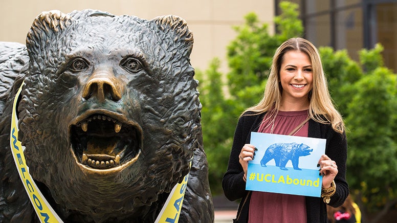 A student holding a #UCLAbound sign smiles as she poses next to The Bruin statue.
